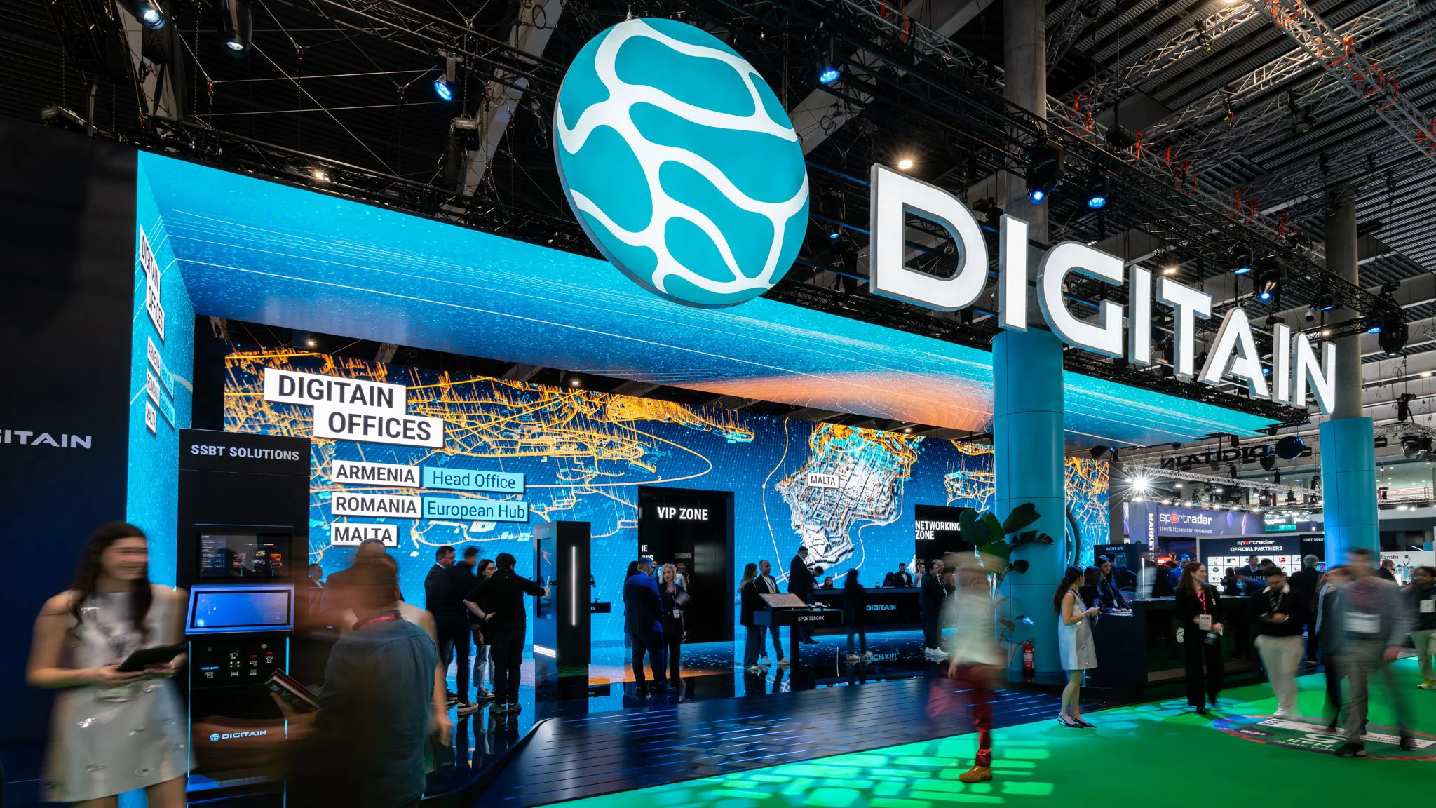 Front view of the Digitain stand entrance with “Digitain Offices” signage and visitors moving through the exhibition hall.